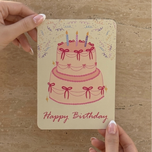 Hand holding a birthday card with a cake illustration and 'Happy Birthday' text on a beige background.