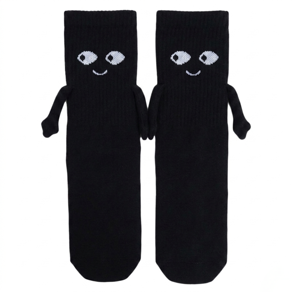 A pair of mid-calf black magnetic hand-holding socks featuring embroidered white cartoon eyes, happy smiles, and small 3D fabric arms magnetically connected in a hand-holding position against a white background.