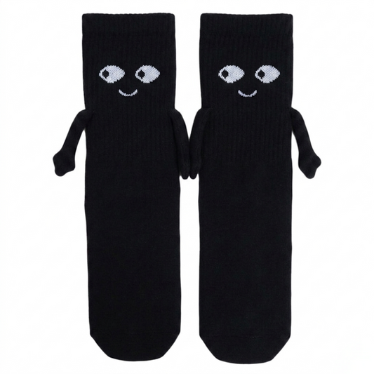 A pair of mid-calf black magnetic hand-holding socks featuring embroidered white cartoon eyes, happy smiles, and small 3D fabric arms magnetically connected in a hand-holding position against a white background.
