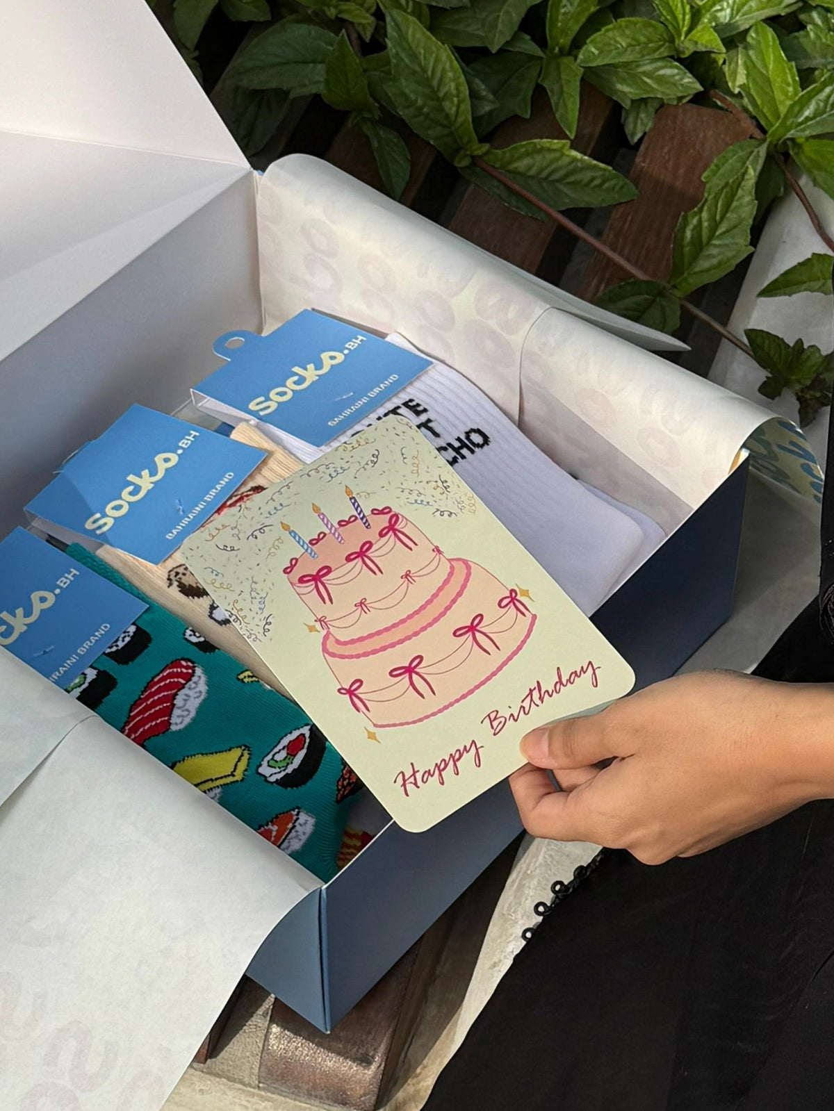 A person's hand holding a pink birthday card with a cake illustration, shown inside an open blue gift box with tissue paper and other cards. Green foliage is visible in the background.